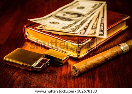 Money with a leather diary and cuban cigar with golden lighter on a mahogany table. Focus on the cuban cigar, image vignetting and hard tones
