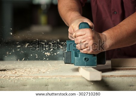 Carpenter working with electric planer on wooden plank in workshop. Royalty-Free Stock Photo #302710280