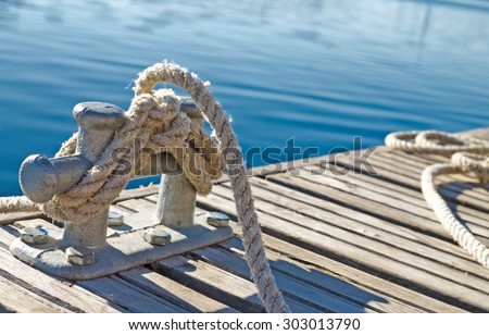 close up of rope tied up on a bitt on wooden dock. blue water in the background