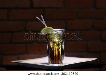 One glass bocal with cold black tea with ice cubes and fresh slice of green yellow citrus lime fruit with two drink straws standing on white plate on brown brick wall background, horizontal picture