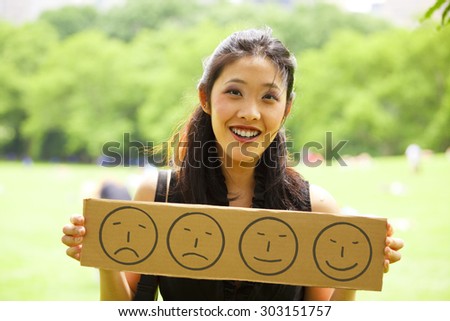 A smiling young woman holding a sign with progressively happier faces. Focus is on her face. Royalty-Free Stock Photo #303151757