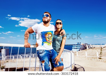 Beautiful young loving couple is standing embracing at pier at blue summer sky background.
