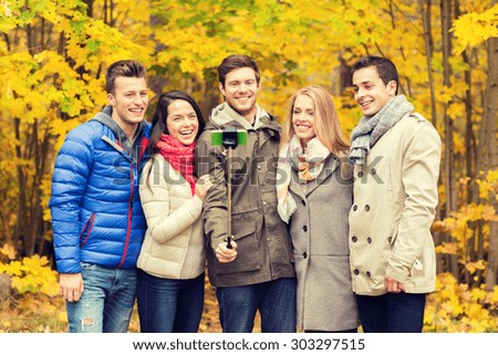 season, people, technology and friendship concept - group of smiling friends with smartphone and selfie stick taking picture in autumn park