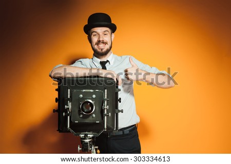 Young  positive  smiling man in hat as photographer with retro camera on an orange background