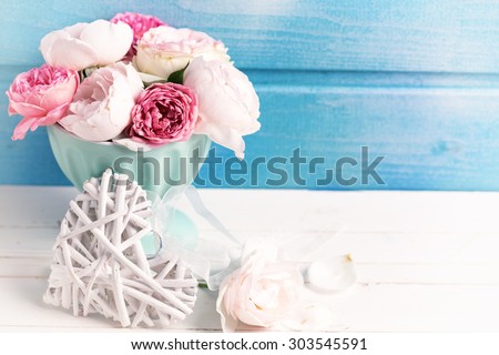 Pastel roses in turquoise vase and decorative heart on white wooden  background against blue wall. Place for text. Selective focus. Toned image.