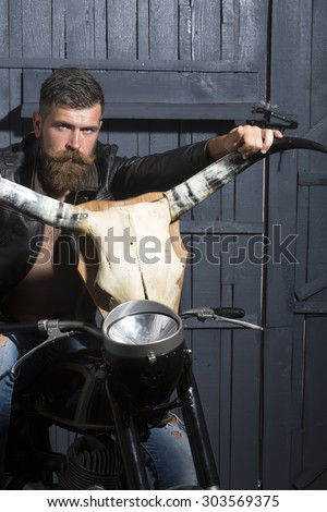 Handsome unshaven male biker in leather jacket sitting on motorcycle in garage with big bone skull antlers of stuffed animal looking forward on wooden wall background, vertical picture