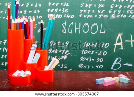 Colorful pencils of red yellow orange violet purple pink green and blue in stationary cup standing on brown school desk on written with white chalk blackboard background, horizontal picture