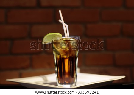 One glass bocal with cold black tea with ice cubes and fresh slice of green yellow citrus lime fruit with two drink straws standing on white plate on brown brick wall background, horizontal picture