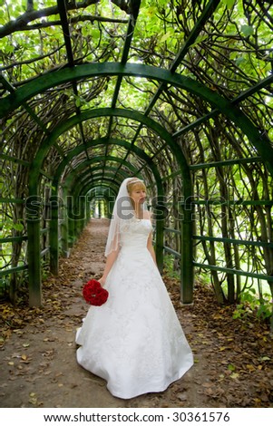 Full-length picture of a bride standing in a green archway and holding a little bouquet of red roses in her hand.