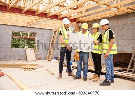 Builder On Building Site Looking At Plans With Apprentices Royalty-Free Stock Photo #303640970