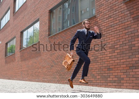 Young businessman with a briefcase and glasses running in a city street on a background of red brick wall Royalty-Free Stock Photo #303649880