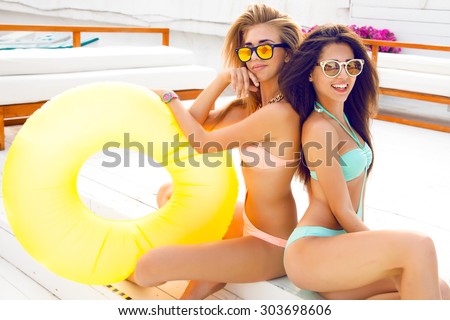 Close up lifestyle fashion portrait of two pretty fresh young brunette and blonde best friends girls, having vacation on the tropical island beach, wearing bikini sunglasses and bright jewelry .
