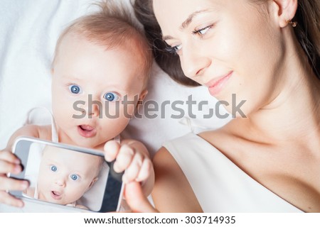Funny baby girl make selfie on mobile phone and lying near her mother on a white bed. Newborn looking at the camera and smiling. Mothercare is most important in baby life Royalty-Free Stock Photo #303714935