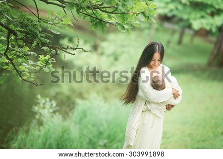 mother and beautiful w?man with long brunette hair in long yellow dress is standing in the park or forest near the trees and holding her little baby girl daughter on her hands hugging and kissing her