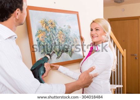 Young man and blonde smiling woman hanging art picture in frame. Focus on woman