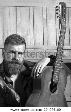 One handsome unshaven musical man with beard and handlebar moustache sitting with acoustic guitar looking forward indoor on wooden wall background black and white, vertical picture