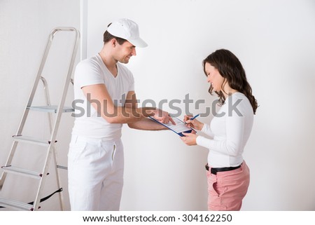 Young Woman Signing Document In Front Of Male Painter At Home