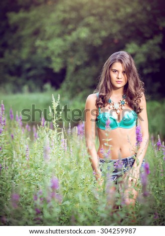 Beautiful young girl standing in high grass and flowers on river bank wearing bra and jeans shorts.