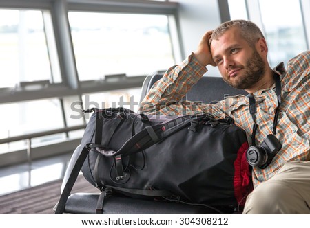 Smiling man at airport. 
Positive bearded passenger sitting airport lounge waiting for his flight casual dress shirt pants large backpack luggage photo camera windows background