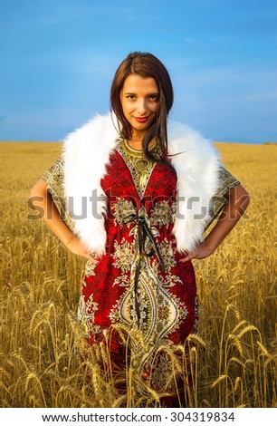  Young woman with ornamental dress and white fur standing on a wheat field with sunset. Natural background.