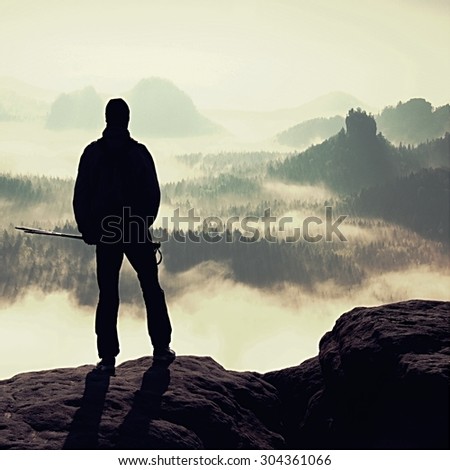 Silhouette of tourist with poles in hand. Hiker stand on rocky view point above misty valley. Sunny spring daybreak in rocky mountains