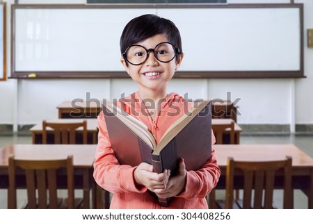 Sweet little girl standing in the classroom while wearing glasses and holding a book, smiling at the camera