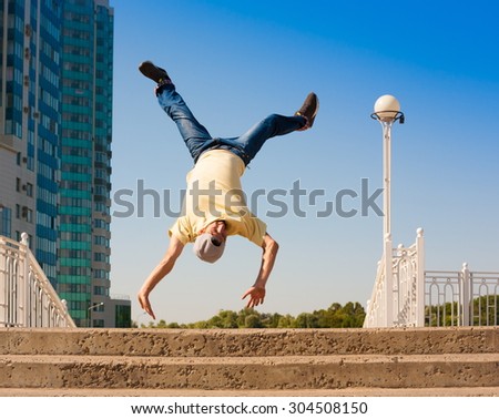 Fun man dancing. Has yellow t-shirt, blue jeans, gray shoes sneakers, slim sport body. Motion on great urban city. Amazing portrait. Sports acrobatic handstand. Fitness concept. Jump creative unique