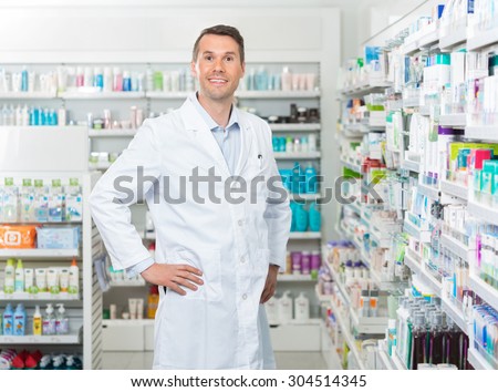 Portrait of confident mid adult male pharmacist standing with hands on hip in pharmacy
