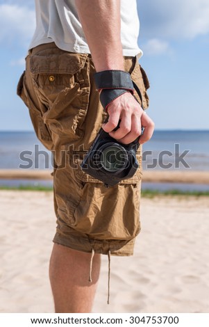 Man (male) dressed in shirt and breeches, standing on sandy shore of sea and holding a camera. Vertical orientation