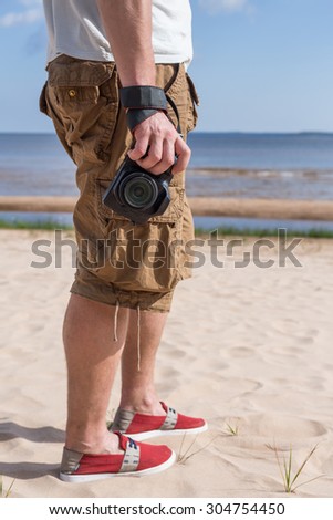 Man (male) dressed in shirt and breeches, red shoes, standing on sandy shore of sea and holding a camera