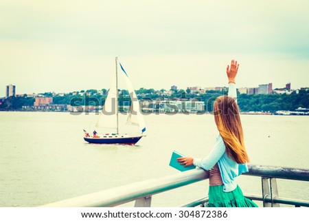 Welcome You. A girl with long blonde hair, wearing cardigan, skirt, holding book, standing by Hudson River in New York, looking at boat in water., raising arm, waving hand. Back view. Instagram effect
