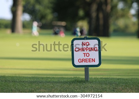 No Chipping to Green Golf Course Sign Positioned near Green, with A Golf Cart and Golfers in the Background.