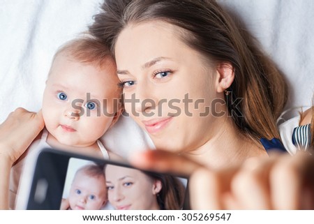 Funny baby girl make selfie on mobile phone and lying near her mother on a white bed. Newborn looking at the camera and smiling. Mothercare is most important in baby life