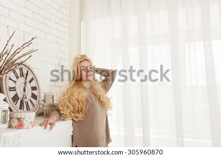 Cute blonde girl in home interior. The girl is wearing a sweater. On a shelf are large clock. The concept of home comfort.