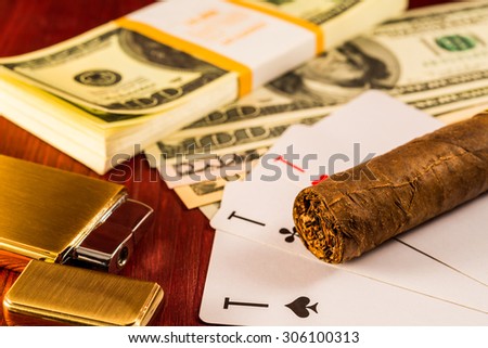 Cuban cigar and golden lighter with playing cards and pack of dollars on the table mahogany. Focus on the cuban cigar, identification cards ace Russian letter