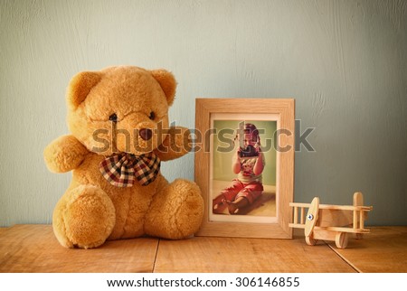 wooden airplane toy and teddy bear over wood table next to photo frame with kid's old photography. retro filtered image
