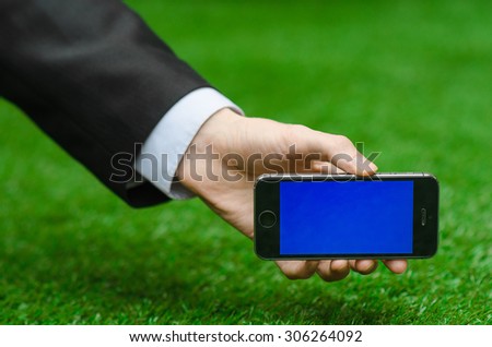 Communication and Business Subject: Hand in a black suit holding a modern phone with blue screen in the background of green grass