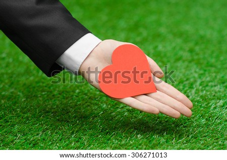 Love and business topic: the hand of man in a black suit holding a card in the shape of a red heart on the background of green grass