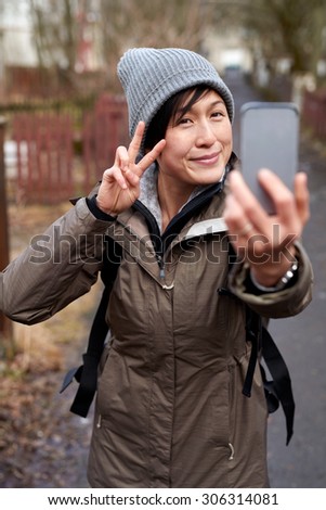 young Asian woman in jacket and hat taking selfie with smartphone while travelling on vacation in Europe