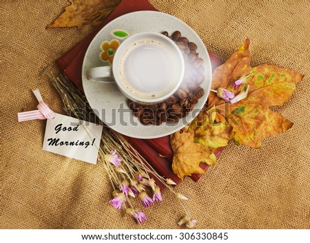 Tag with words good morning and the cup of coffee lying on the books with dry yellow maple leaves and everlasting flowers on sackcloth background