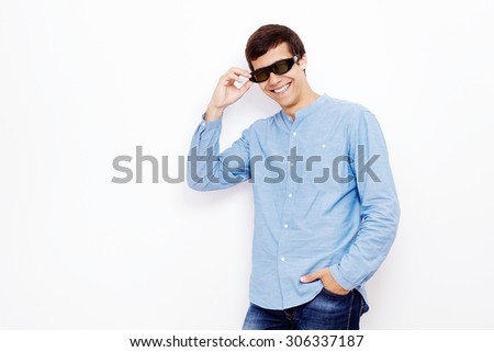 Young hispanic man wearing jeans shirt standing with hand on his 3D TV LCD shutter glasses and smiling against white wall - 3D film concept