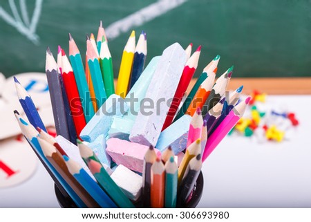 Colorful pencils of red yellow orange violet purple pink green and blue in stationary cup on school desk with white sheet of paper on written with chalk blackboard background, horizontal picture