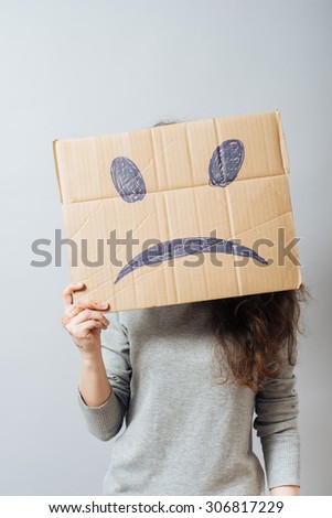 Young woman holding cardboard sad smiley. On a gray background.