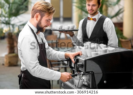Handsome barista in uniform making coffee standing with waiter in classical renaissance cafe patio