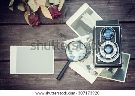 The old blank photo is lying on a wooden desk  in the left  edge of the photo. On the right side of the photo is laying magnifier and old camera on an old photos. Dried red rose is in the upper part. 