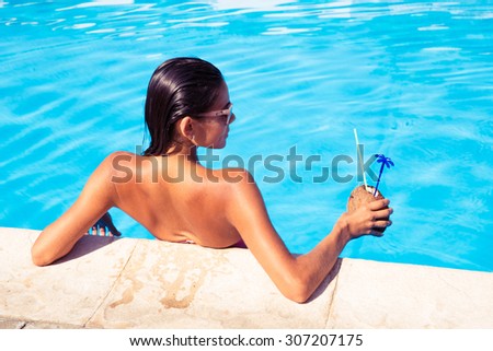 Back view portrait of a young woman standing in swim pool and holding cocktail in coconut outdoors