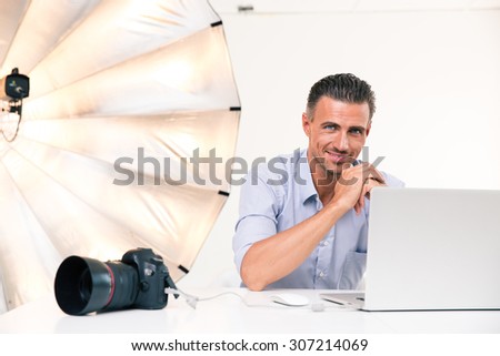 Portrait of a smiling photographer using laptop at his workplace and looking at camera