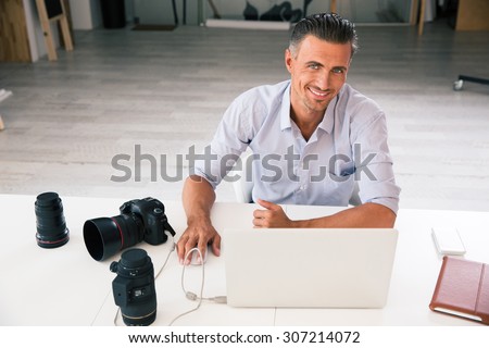 Portrait of a happy photographer using laptop at his workplace and looking at camera