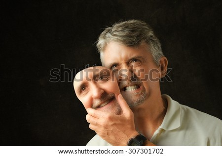 An angry, emotional, depressed, bipolar disorder man is revealing his true self as he takes off a fake smile happiness mask that looks exactly like his face.  Royalty-Free Stock Photo #307301702