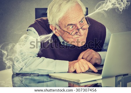 Stressed elderly old man using computer blowing steam from ears. Frustrated guy sitting at table working on laptop isolated on gray wall background. Senior people and technology concept  Royalty-Free Stock Photo #307307456
